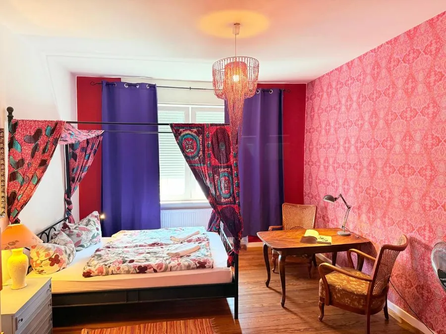 Doppelzimmer mit rosa gemusterter Tapete, Doppelbett, Schreibtisch und Balkontür